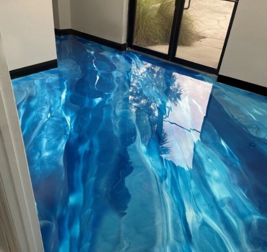 Room with a glossy, blue epoxy floor resembling water over concrete, reflecting a window and outdoor scenery.