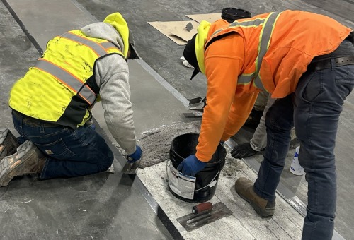 Two workers in high-visibility gear repairing a concrete floor, using tools and a bucket of materials.