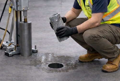 Worker in safety gear examines a cylindrical concrete core sample from a drilled hole in the floor, surrounded by water.