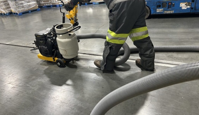 Worker using industrial equipment on smooth concrete floor, wearing reflective work gear. Large hoses connected to machine.