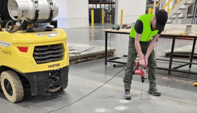 Worker in a neon vest drilling into a concrete floor in a warehouse, with a yellow forklift nearby and construction materials on a table.