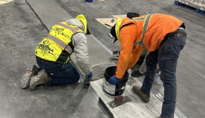 Workers in safety gear repair a concrete floor, kneeling next to a bucket and tools.