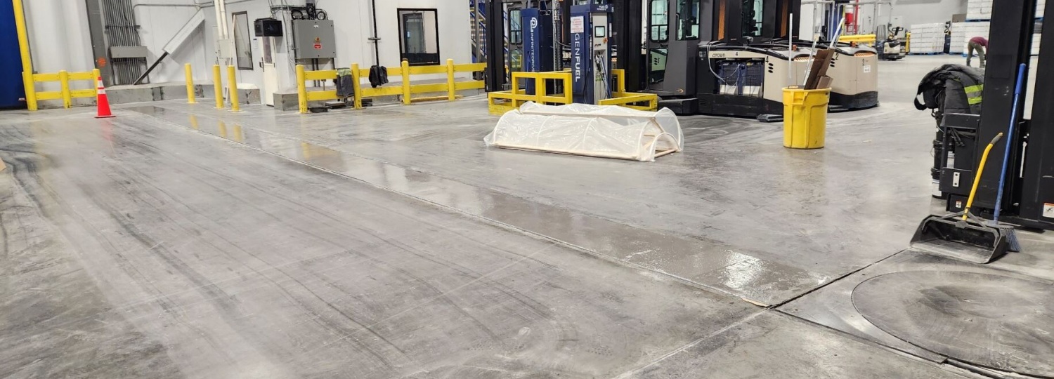 Warehouse interior with polished concrete floor, yellow barriers, a traffic cone, forklifts, and cleaning equipment visible.