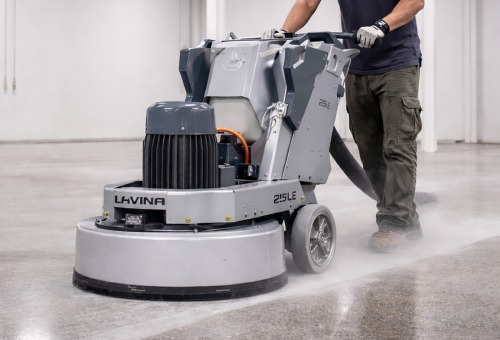 Worker operating a Lavina 215 LE machine, smoothing a concrete floor in an indoor setting. Dust is visible around the machine.