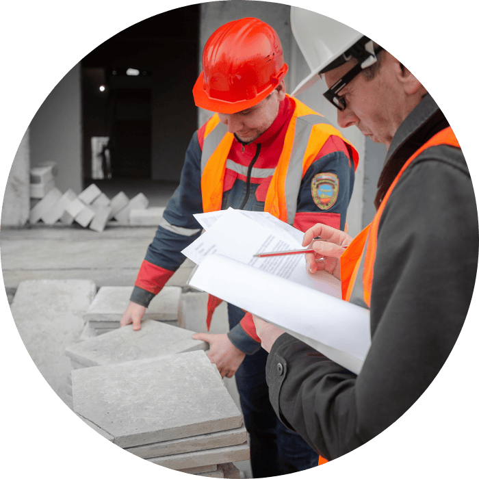 Construction workers in safety gear examine concrete slabs, holding documents and a pencil at a building site.