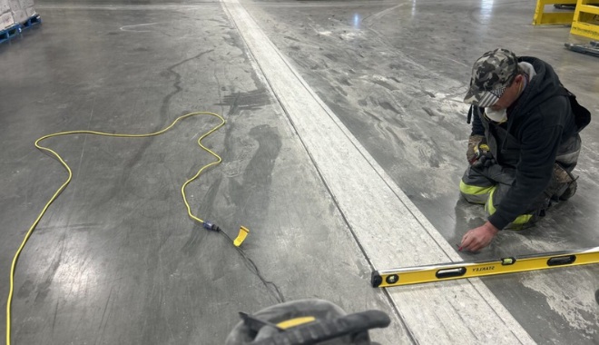 Worker kneeling on concrete floor, using a spirit level to check for evenness. An extension cord lies nearby, indicating ongoing work.