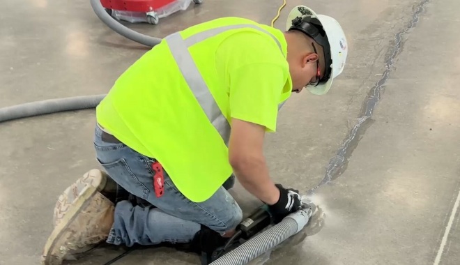 Worker in a neon vest and hard hat kneels on a concrete floor, operating machinery to repair a long crack.