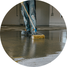 Person smoothing wet concrete floor with a large broom in an unfinished room.