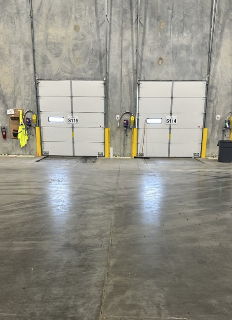 Two garage doors labeled S115 and S114 in a concrete warehouse interior, with safety equipment and yellow bollards.
