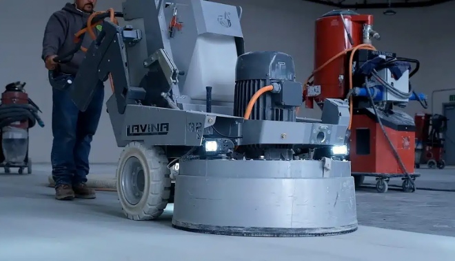 Worker operating a large concrete polishing machine in an industrial setting, with equipment visible in the background.