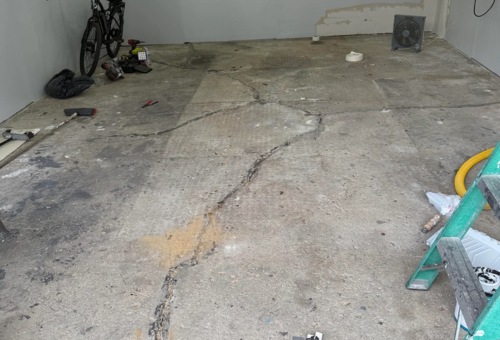 Garage with cracked concrete floor, scattered tools, a fan, ladder, and bicycle leaning against the wall.