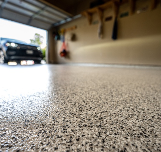 Smooth, speckled concrete garage floor with open door showing a car parked outside, shelves on the right wall.