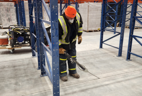 Worker in safety gear using a tool to cut concrete slab in industrial warehouse with blue metal shelving and stacked pallets.