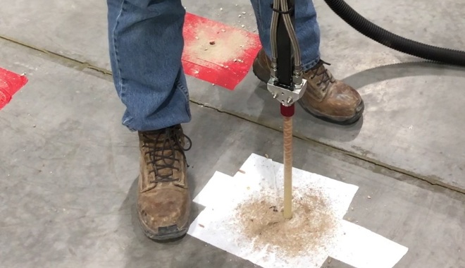 Worker drilling into a concrete floor with a machine, surrounded by debris and a paper sheet. Dust collector attached to reduce mess.