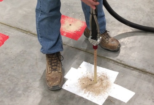 Person drilling into concrete floor, using a tool with a vacuum attachment to minimize dust, wearing jeans and work boots.