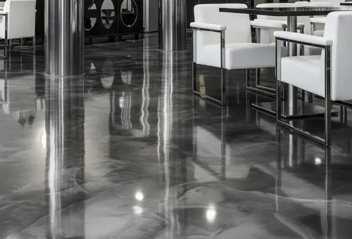 Glossy polished concrete floor in a modern space with white chairs and metallic table legs reflecting on the surface.
