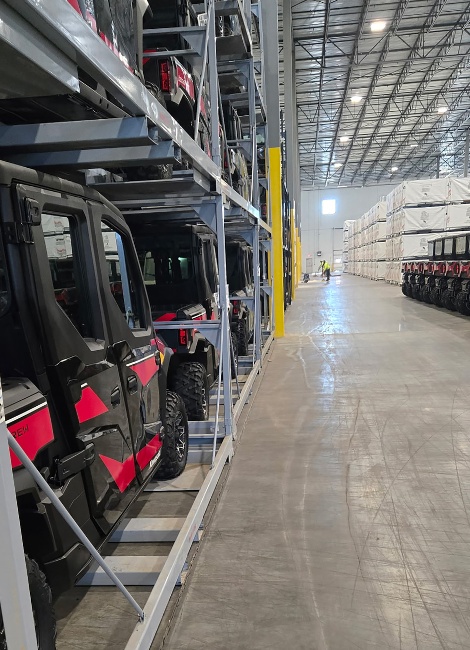 Warehoused vehicles on stacked metal racks alongside neatly arranged, wrapped packages on a polished concrete floor. High ceiling warehouse.