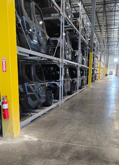 Warehouse interior with stacked utility vehicles on metal racks, concrete floor, yellow pillars, and a fire extinguisher attached to a pillar.