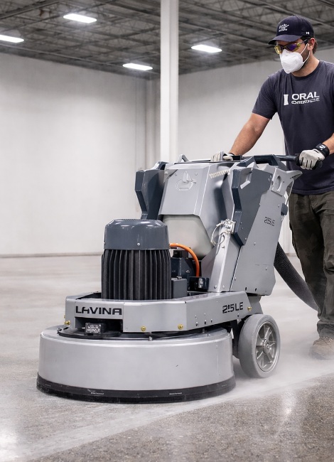 Worker polishing smooth concrete floor with a large, gray machine in an industrial setting, wearing safety gear.