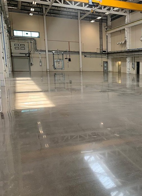 Large industrial interior with polished concrete floors, overhead piping, and a yellow crane beam. Natural light streams through high windows.