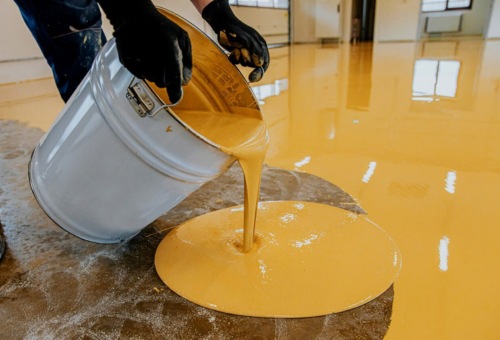 Yellow epoxy poured from a container onto a concrete floor, creating a smooth, glossy surface. Person holding the container wears black gloves.