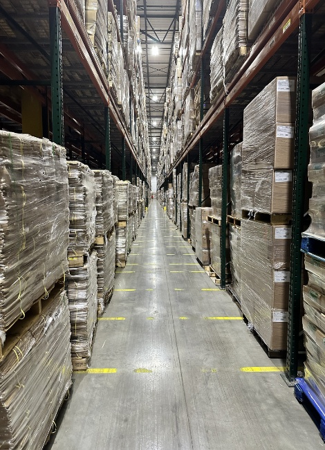 Warehouse aisle with tall shelves stacked with goods on both sides, clear overhead lighting, and smooth concrete flooring.