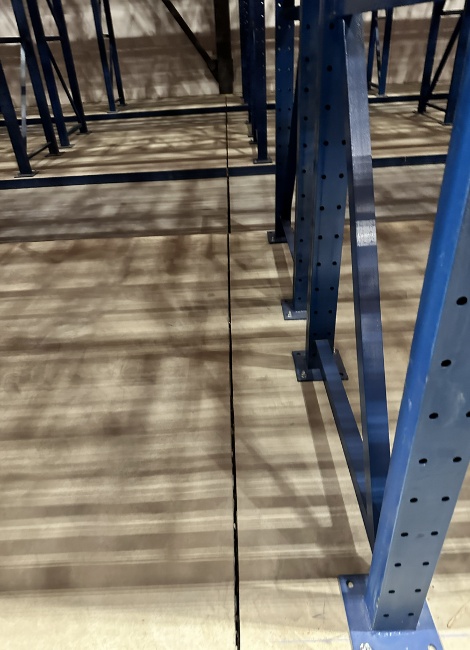 Warehouse aisle with blue metal shelving on a concrete floor, showing shadows and bolt holes on support beams.
