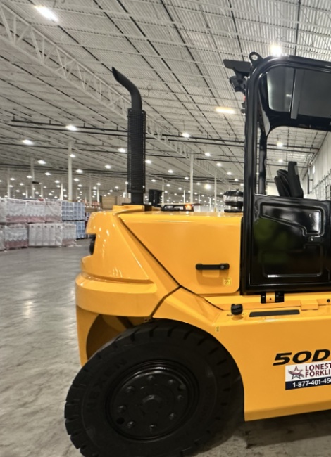Yellow forklift on a concrete floor in a spacious warehouse with stacked goods and high ceilings in the background.
