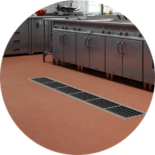 Commercial kitchen with stainless steel appliances, orange flooring, and a grated drain. Cabinets sit against a concrete wall.