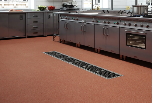 Commercial kitchen with stainless steel appliances, red flooring, and a central floor drain. Concrete walls visible in the background.