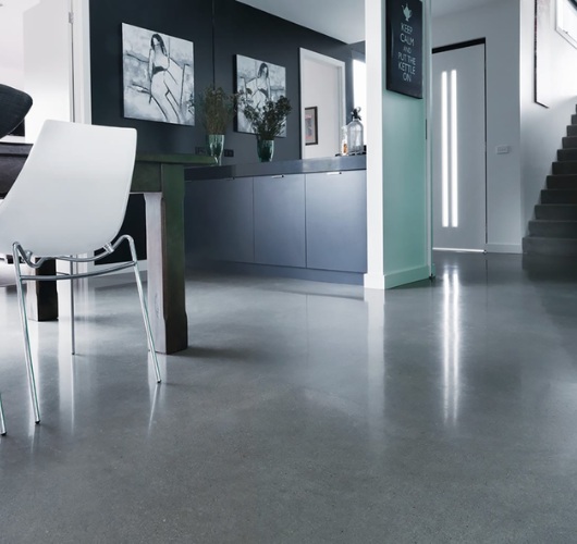 Modern kitchen with sleek concrete floor, white chair, dark cabinetry, art on walls, and a staircase to the right.