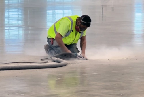 Worker in hi-vis vest kneeling, smoothing concrete floor with a tool, surrounded by dust inside a large, bright indoor space.