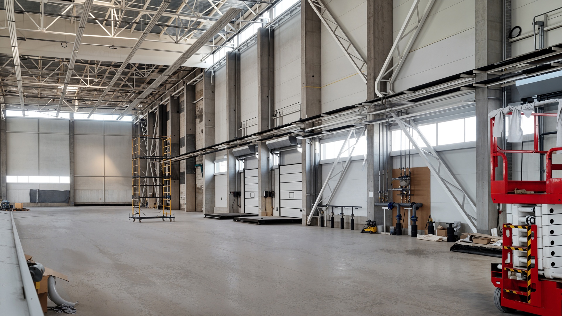 Spacious industrial warehouse with high ceilings, exposed concrete walls, scaffolding, and a red lift on the right.